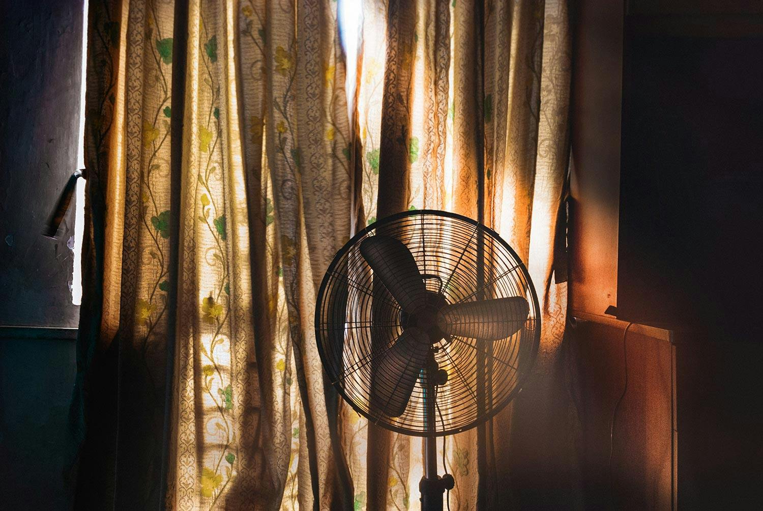 An electric fan sits by gold window curtains with a green botanical print. Rays of sunlight pass through the window.