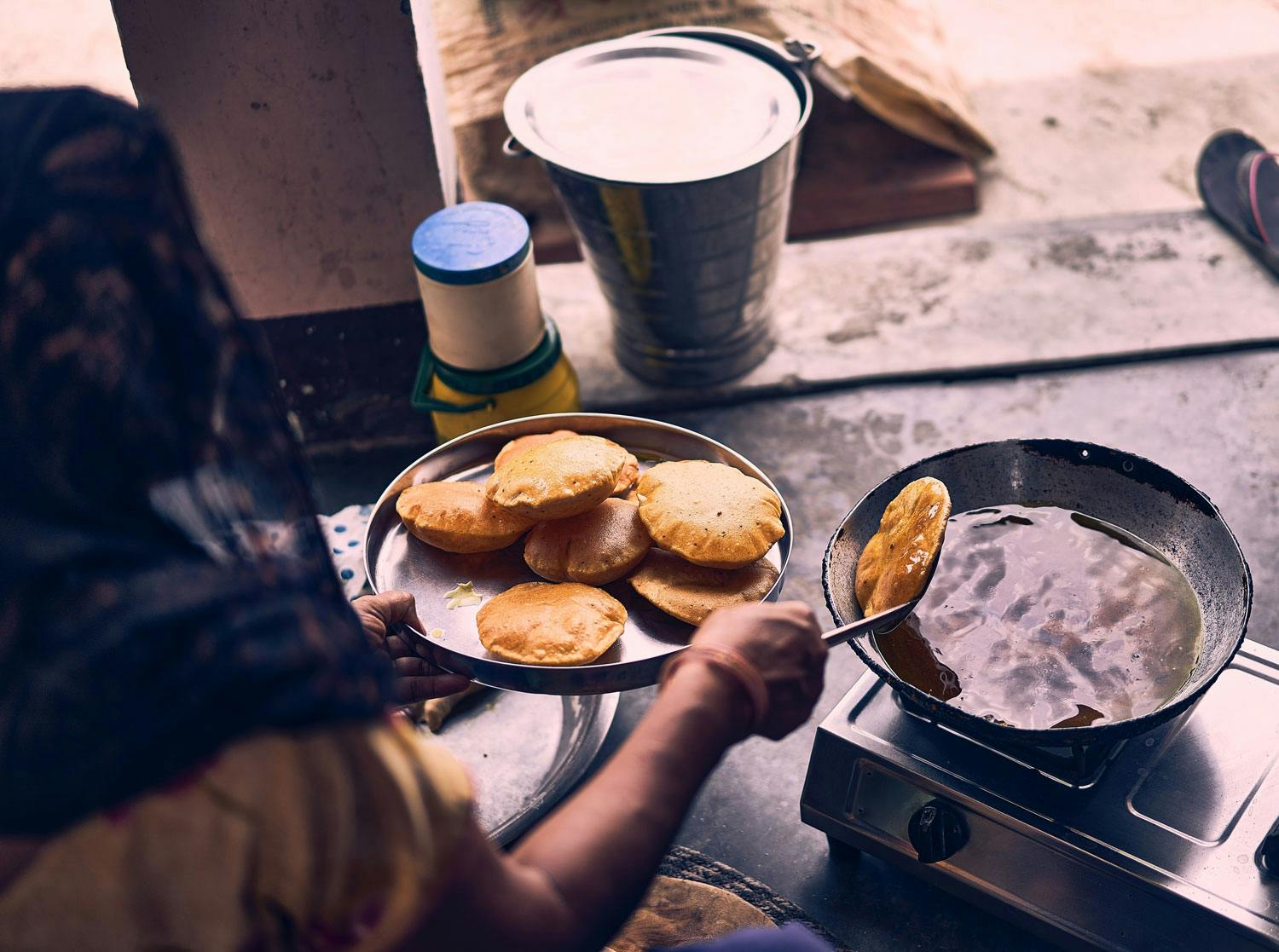 A woman scoops pooris, disks of puffed, fried dough, from a pot of oil with a steel ladle.