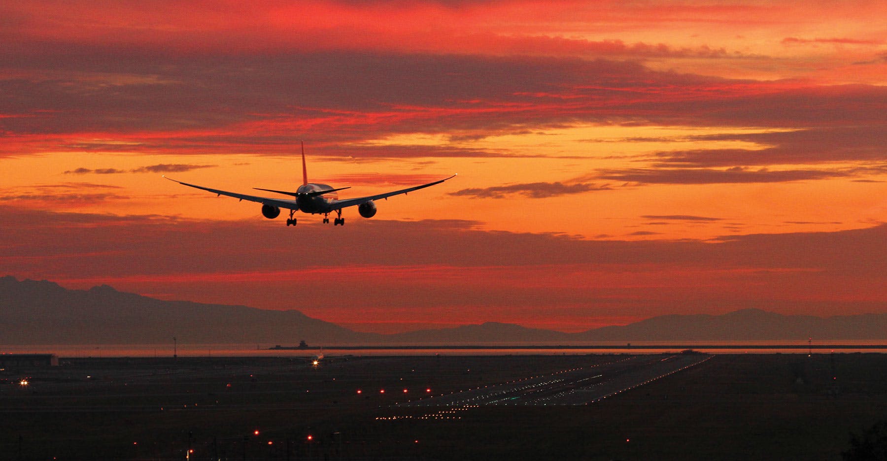 An airplane flying toward a deep orange sunset, preparing to land on a runway lit up with sparkling lights.