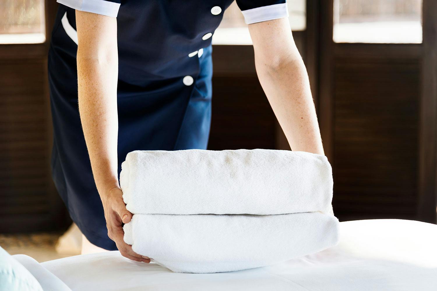 A hotel housekeeping staff member wearing a dark blue uniform places two folded white towels onto a white bedspread.