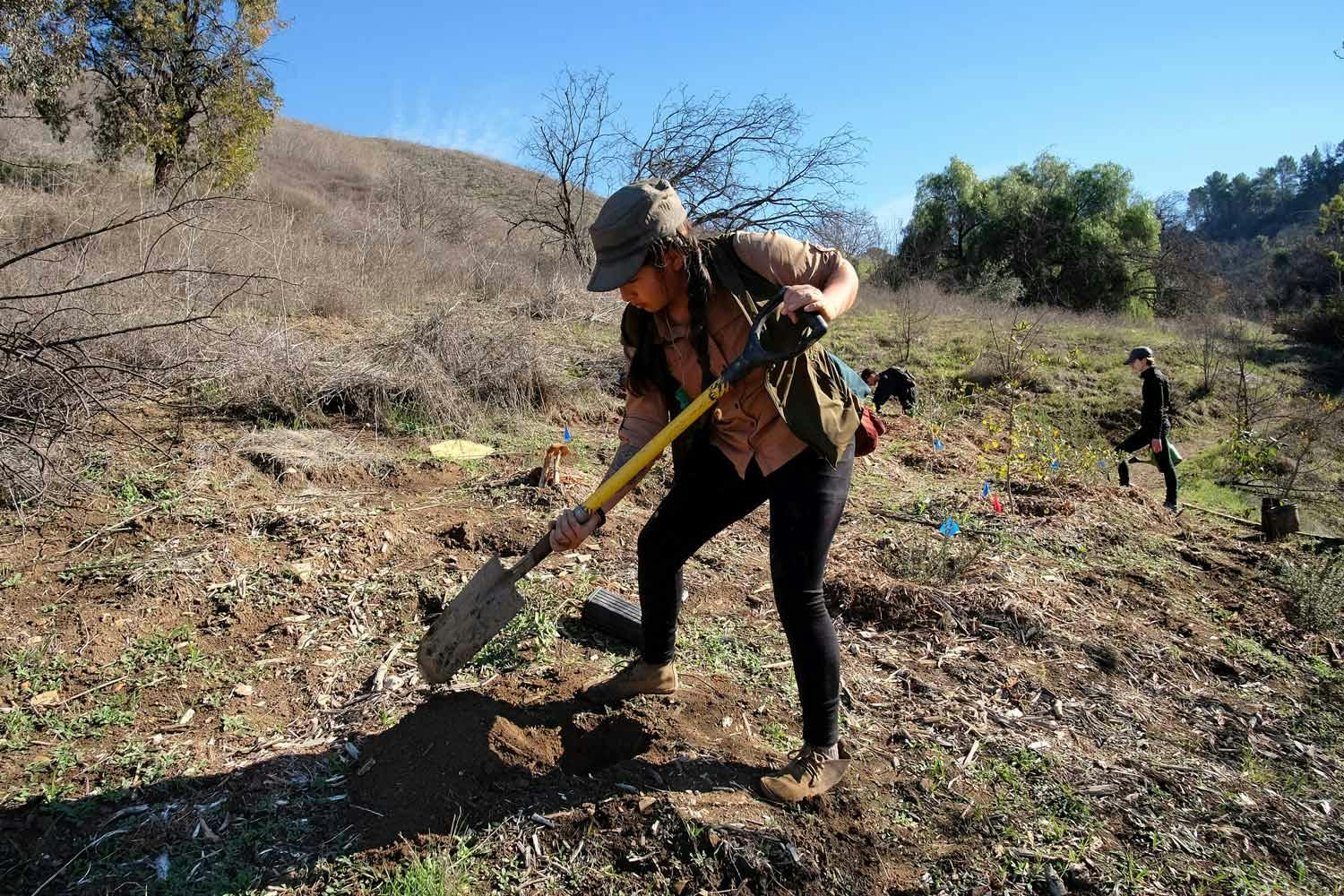 A young woman wearing a cap, vest, and black pants uses a shovel to dig soil on a grassy hillside in Los Angeles.