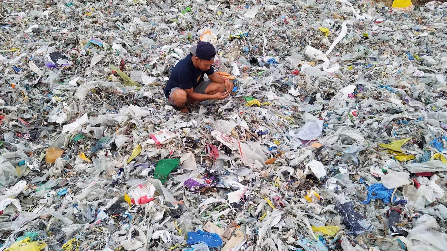 A man crouching amidst a massive pile of plastic garbage.