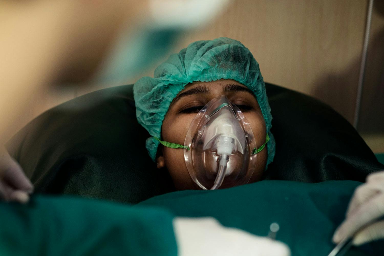 A person lying on a stretcher wearing a green hair net and an oxygen mask, eyes closed.