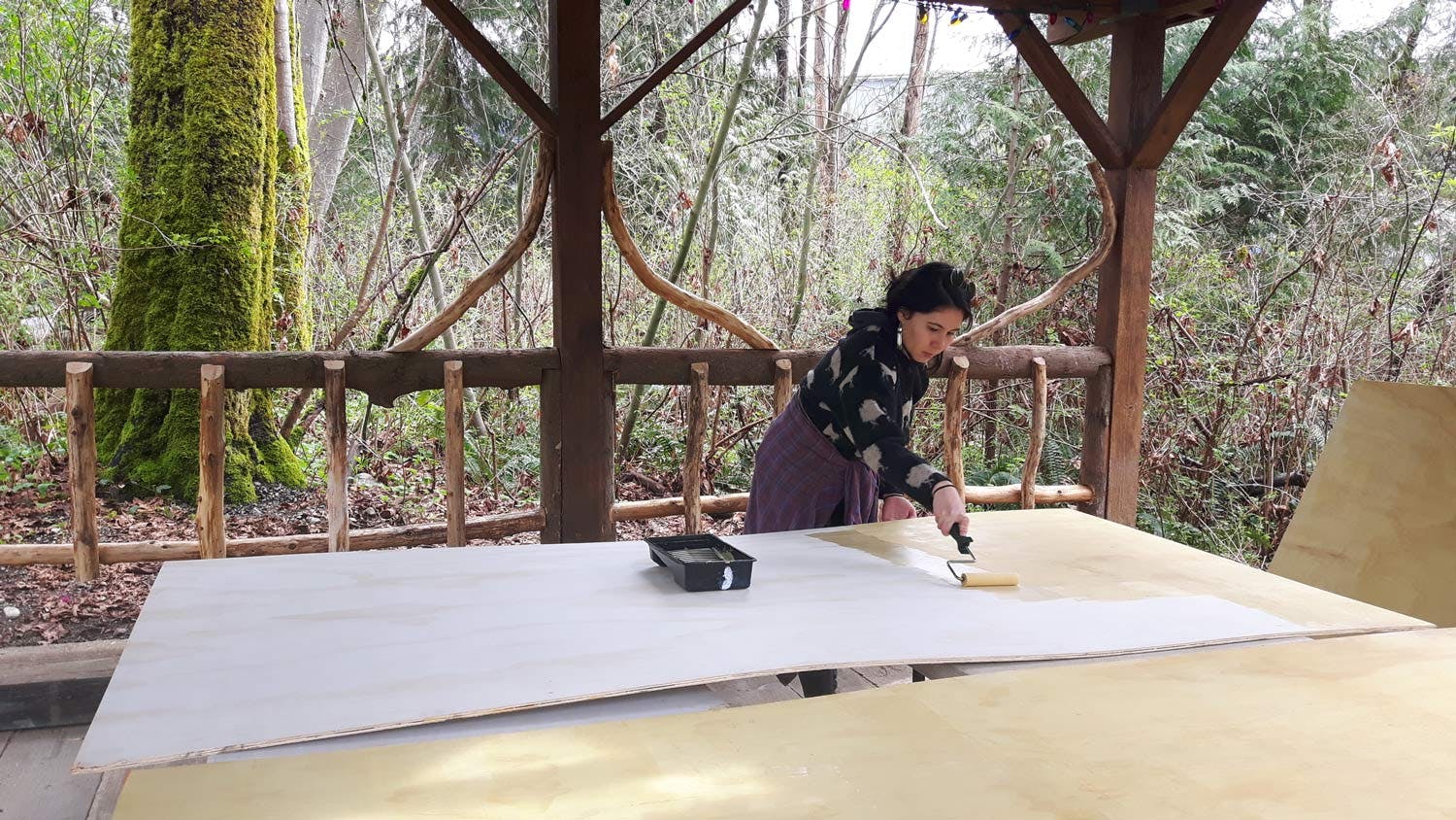 Ocean Hyland rolls primer onto large sheets of plywood under a wooden gazebo in a forest.