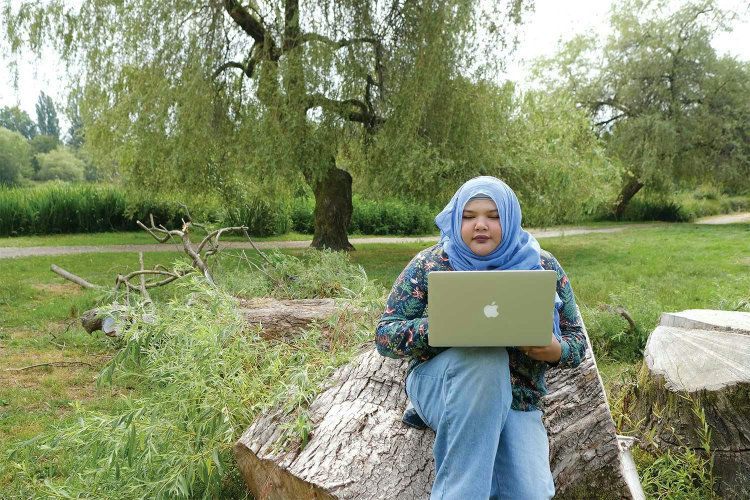 Aliya Abbas sits on a stump at a park. She wears a blue hijab, a floral print shirt and jeans, and works on her laptop.