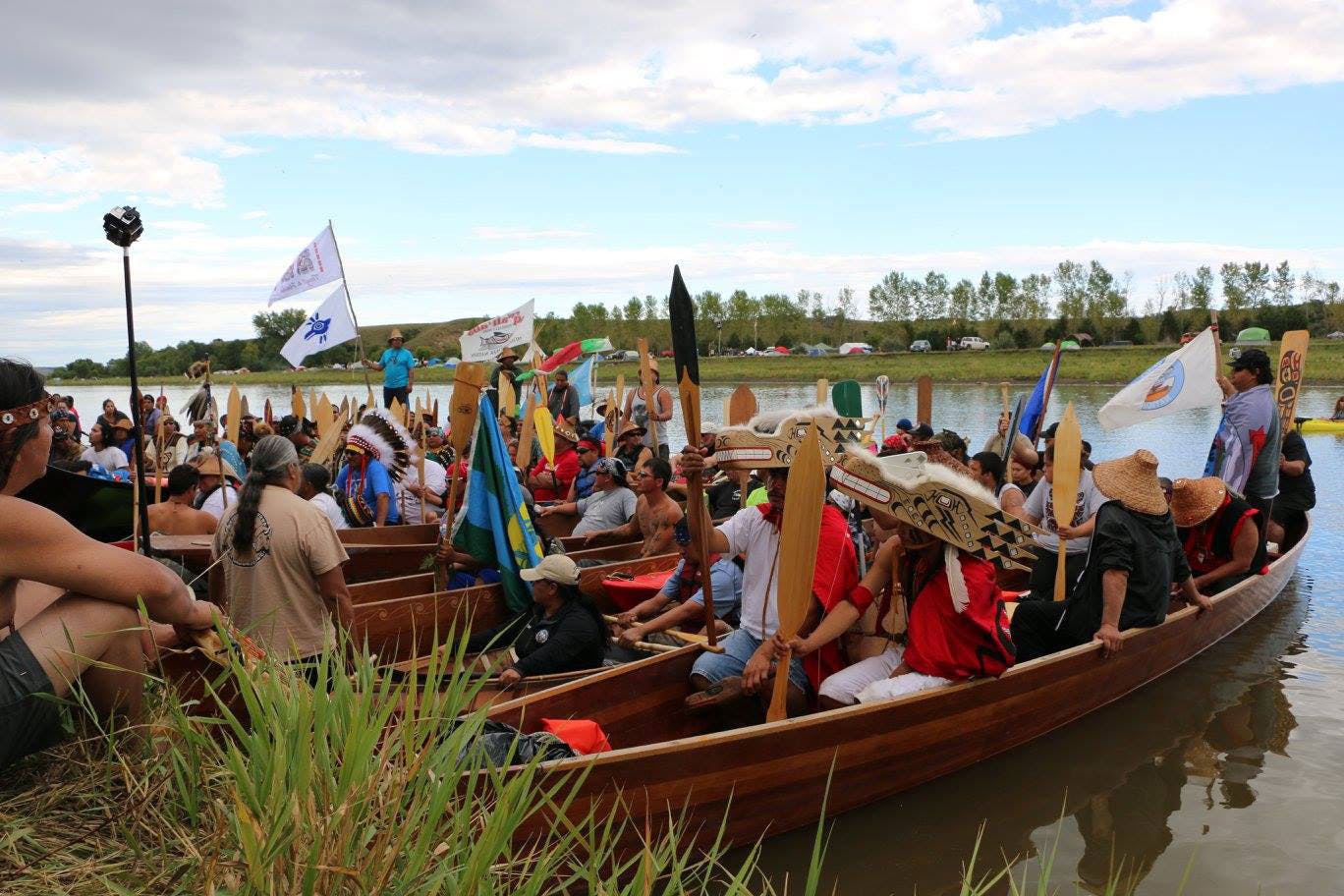 Indigenous activists, holding flags and oars, make a stirring entrance at Standing Rock as they arrive in a line of canoes.