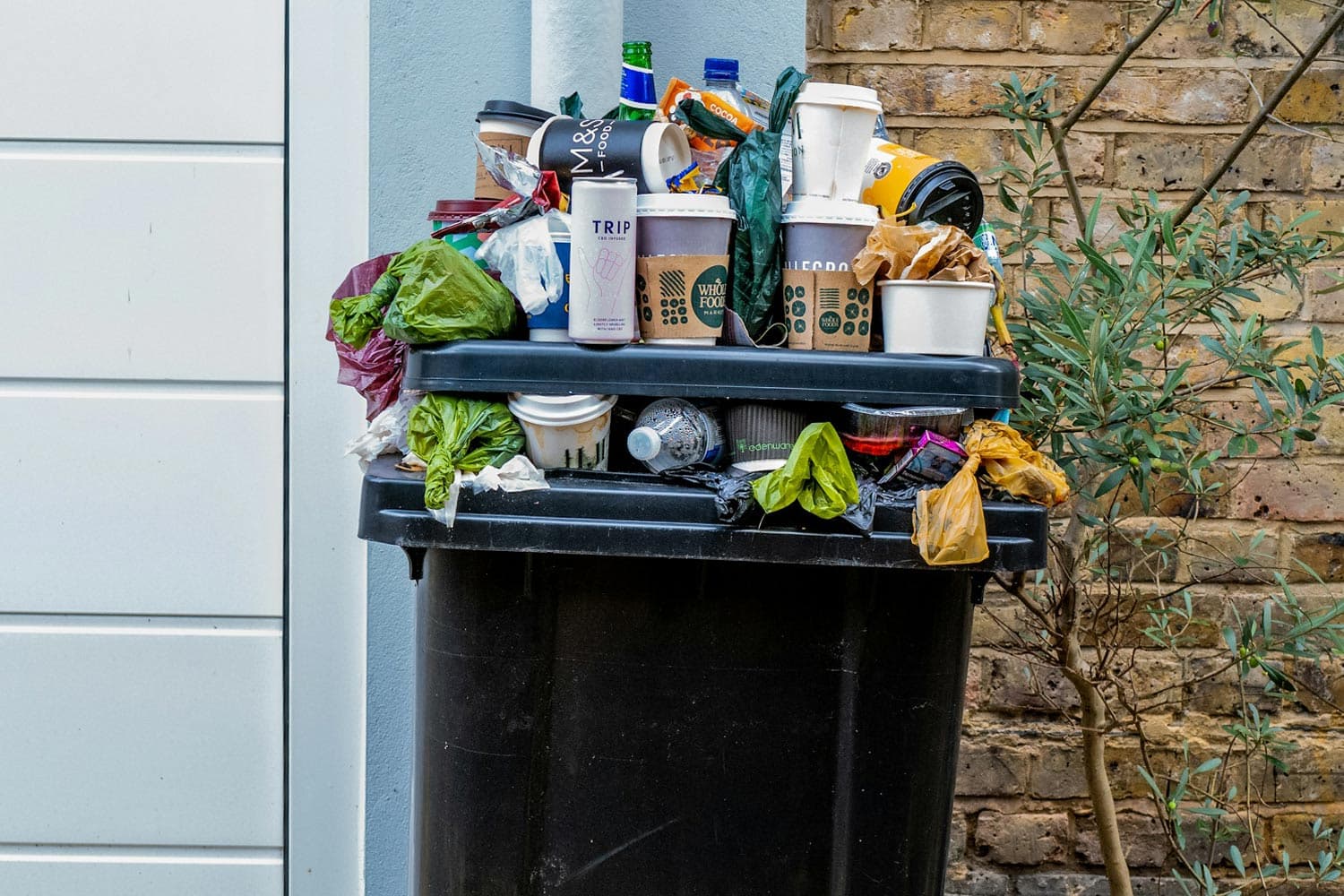 A black plastic garbage bin overflows with plastic bags, bottles, and coffee cups.