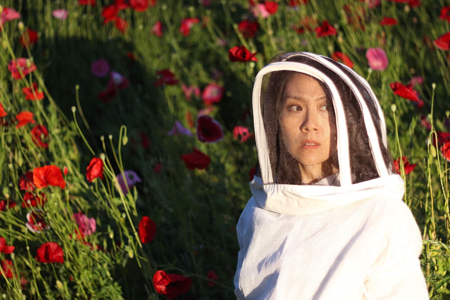 Woman with tan skin and dark hair wears a white beekeeping suit and stands in bright sunshine casting a shadow on a field of red and pink poppies.