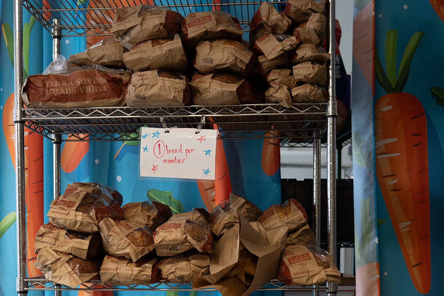 Paper bags of bread fill a metal rack above a handwritten sign reading “1 bread per member.”